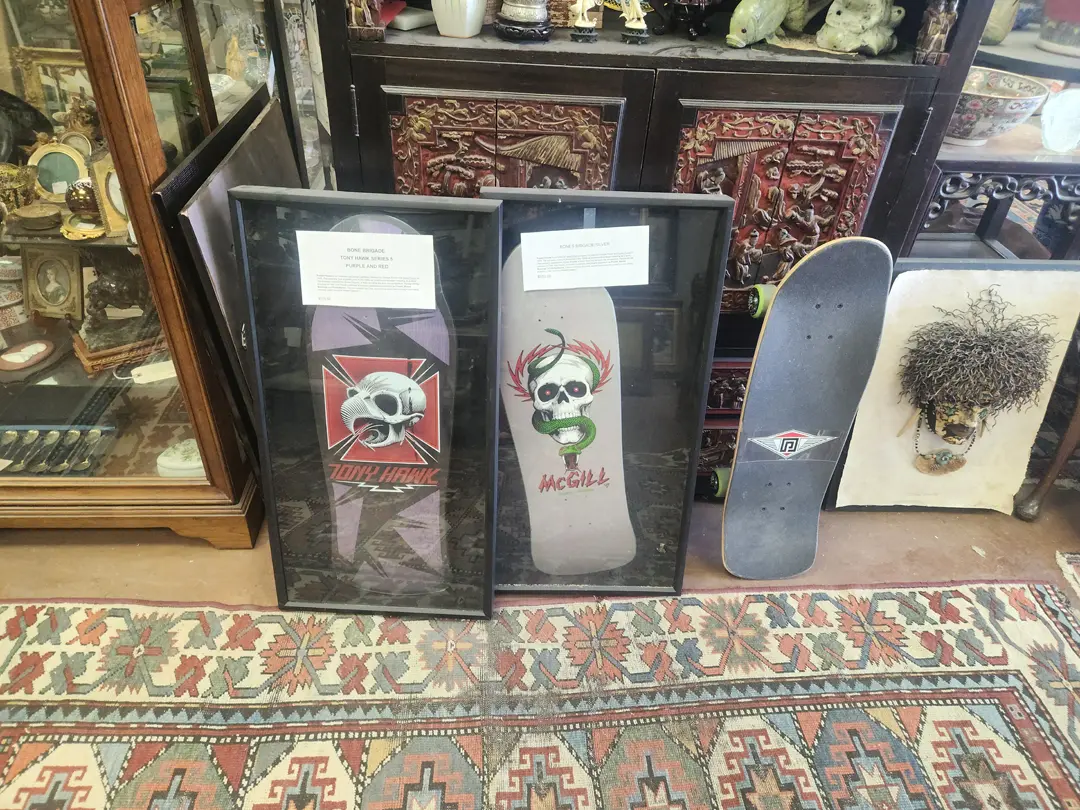 Framed artwork with skull designs sits next to a skateboard and a wall-mounted decoration, displayed on an ornate rug alongside vintage oil paintings in a room filled with various Southwestern items at Red Coyote Trading Post in Ft. Mohave, AZ.