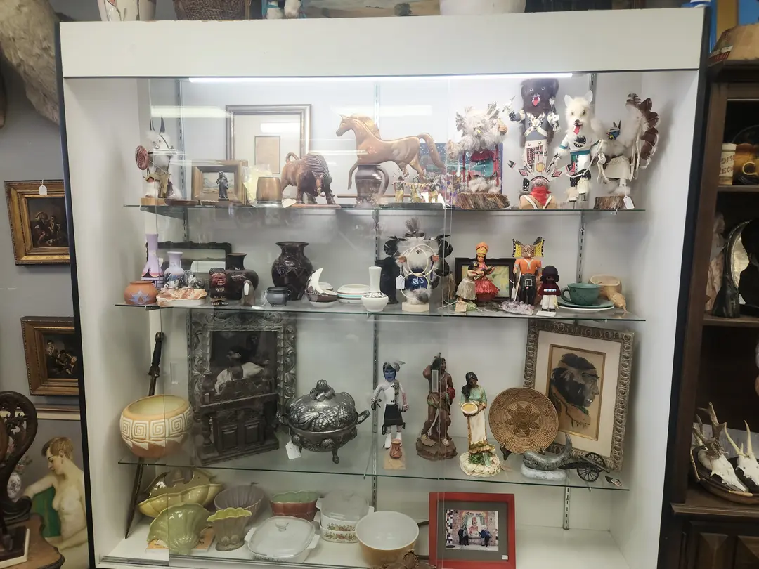 A stunning display cabinet at Red Coyote Trading Post in Ft. Mohave, AZ is filled with an eclectic array of collectibles, including handcrafted figurines, Southwestern vases, and unique decorative items. These treasures are beautifully arranged on shelves set against a mirrored back. Adding to the charm and character of the ensemble are vintage oil paintings that reflect the spirit of the Southwest.