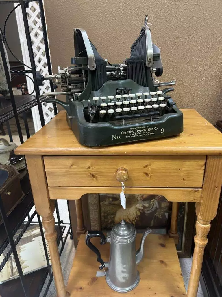An antique typewriter from Red Coyote Trading Post in Ft. Mohave, AZ, sits on a rustic wooden side table with a vintage coffee pot beneath.