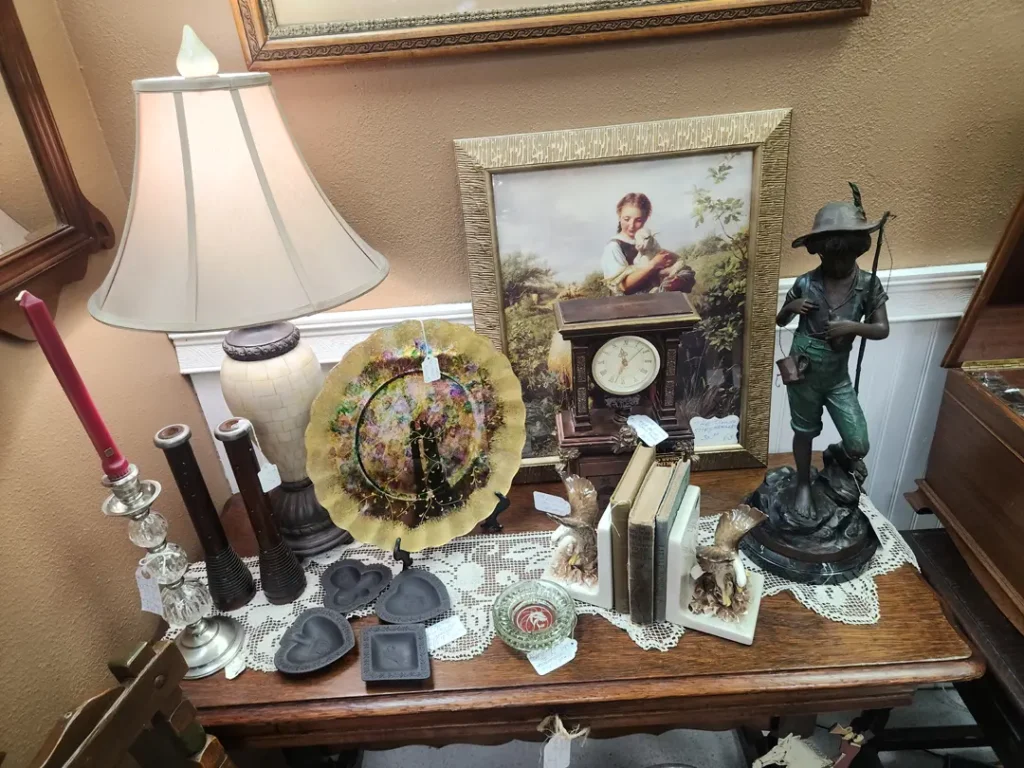 A table displays a variety of items, including a lamp, picture frame, decorative plate, candles, statue, and books. Some items are on a lace doily. A framed painting leans against the wall.