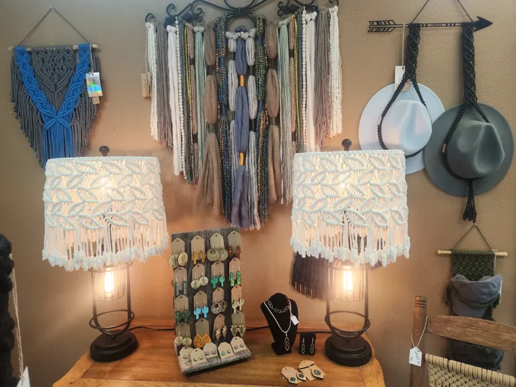 Table with two lamps featuring crochet shades, assorted handmade jewelry displayed on a stand, and wall-mounted hats and macramé decorations in a cozy craft shop.