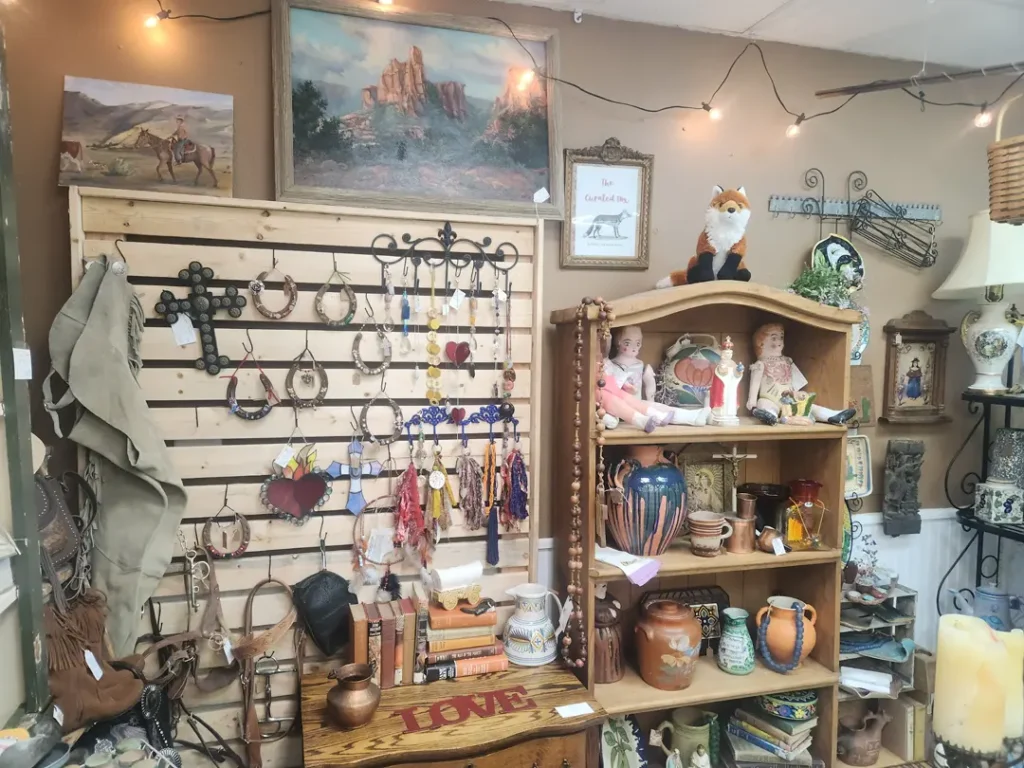 A vintage shop display with ceramics, books, horse decor, and paintings. Shelves and walls are filled with various items, including dolls, a plush fox, and decorative lights.