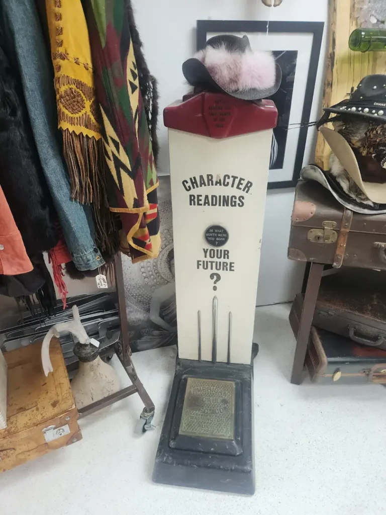 Vintage character reading machine, with a hat on top, surrounded by eclectic items including clothing, suitcases, and a guitar.