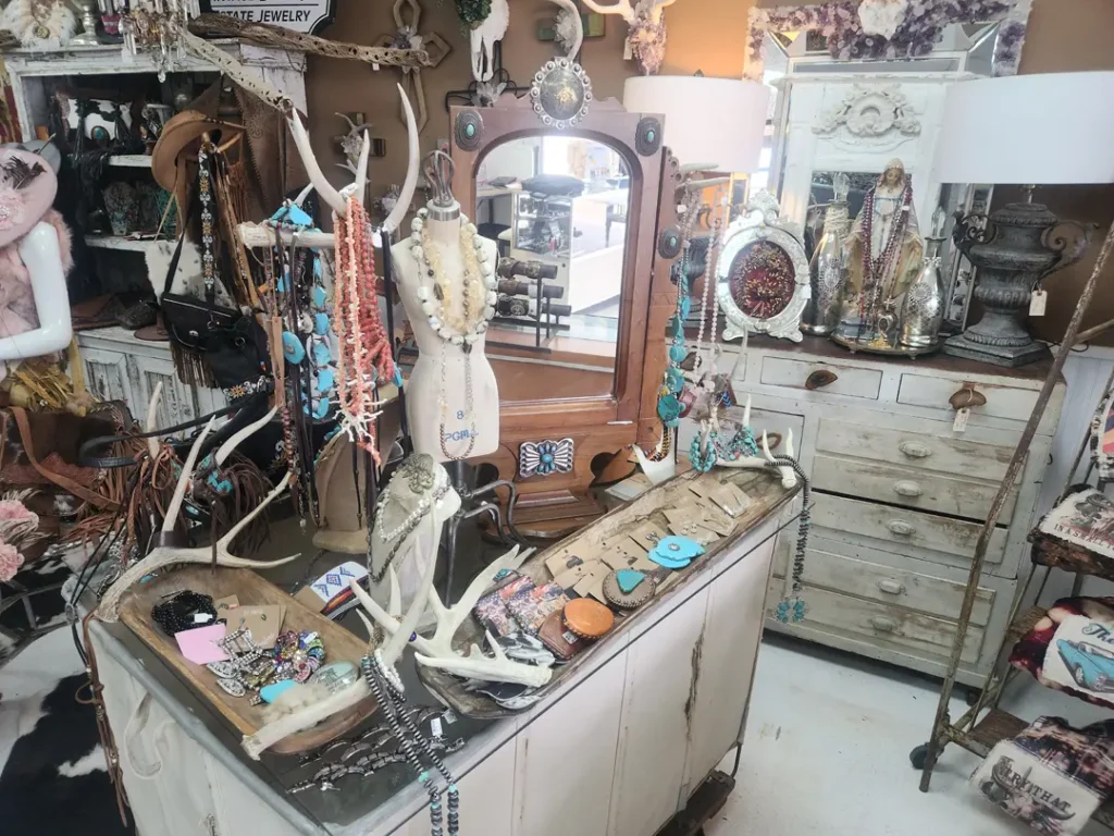 A jewelry boutique display with necklaces, bracelets, and earrings on a wooden dresser and mannequin. A mirror and various decorative items are in the background.