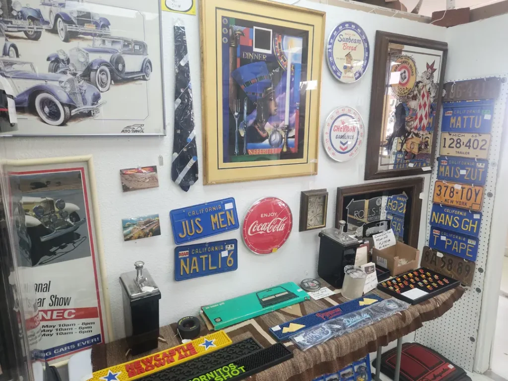 Red Coyote Trading Post in Ft. Mohave, AZ presents an engaging wall display featuring vintage car memorabilia that's a treasure trove for enthusiasts. The collection includes framed classic car photos, authentic vintage license plates, automotive-themed ties, an iconic Coca-Cola sign, and meticulously crafted scale car models. On the accompanying table sits an array of smaller collectibles and a retro typewriter, perfect for those with a passion for history and nostalgia.